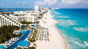 Aerial view of a vibrant beach with resort buildings, turquoise waters, and sun loungers along the sandy shoreline under a clear sky.
