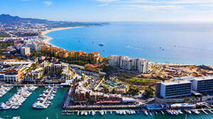 Aerial view of a vibrant coastal city with a marina, luxury boats, and a sandy beach under a clear blue sky.