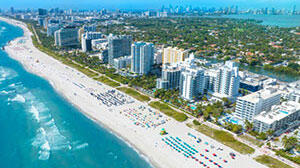 Aerial view of a sunny beach with white sand, colorful umbrellas, and tall buildings lining the coast, against a backdrop of blue ocean.