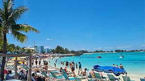 Crowded beach scene with sunbathers and colorful umbrellas, clear turquoise water, and palm trees under a bright blue sky.