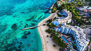 Aerial view of a tropical resort along a sandy beach, with clear turquoise waters and lush greenery surrounding the area.