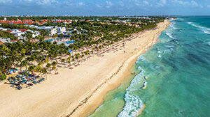 Aerial view of a sandy beach lined with palm trees, colorful resorts, and turquoise waves under a bright blue sky.